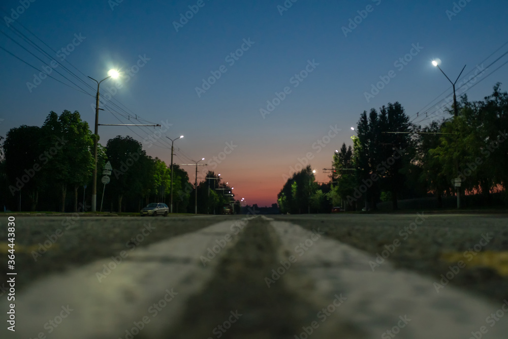 Paved road on the outskirts of the city at sunset, on the sides of green trees grow, lights are burning