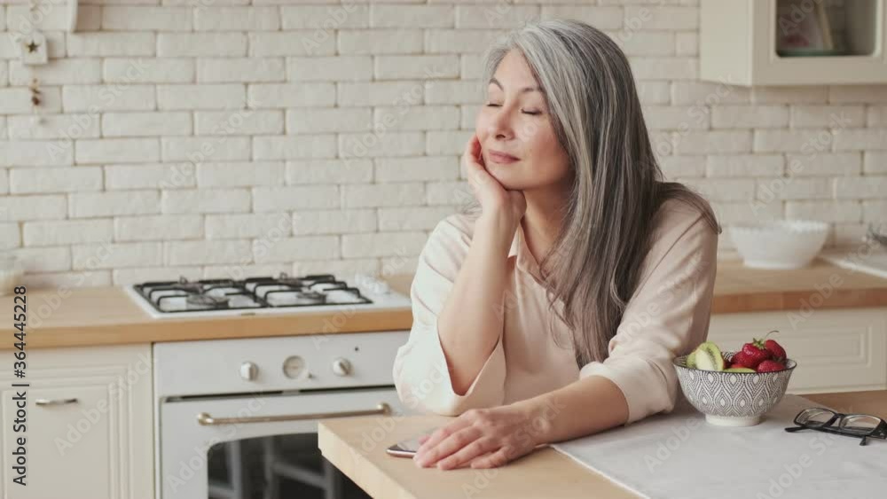 A dreaming relaxed mature woman is sitting at the table in the kitchen and looking to the side at home A dreaming relaxed mature woman is sitting at the table in the kitchen and looking to the side at home