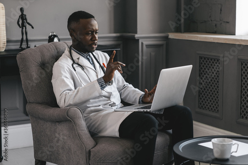 dark-skinned doctor talking on skype zoom with laptop waving photo