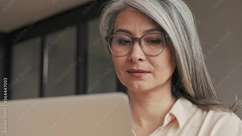 A close-up view of a calm mature woman in eyeglasses is using her laptop at home A close-up view of a calm mature woman in eyeglasses is using her laptop at home