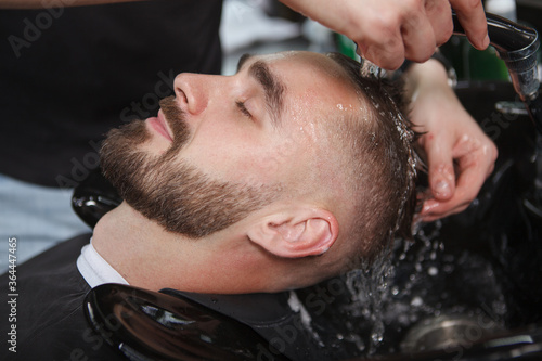 Happy handsome bearded man relaxing while professional barber washes his hair after shaving and giving haircut
