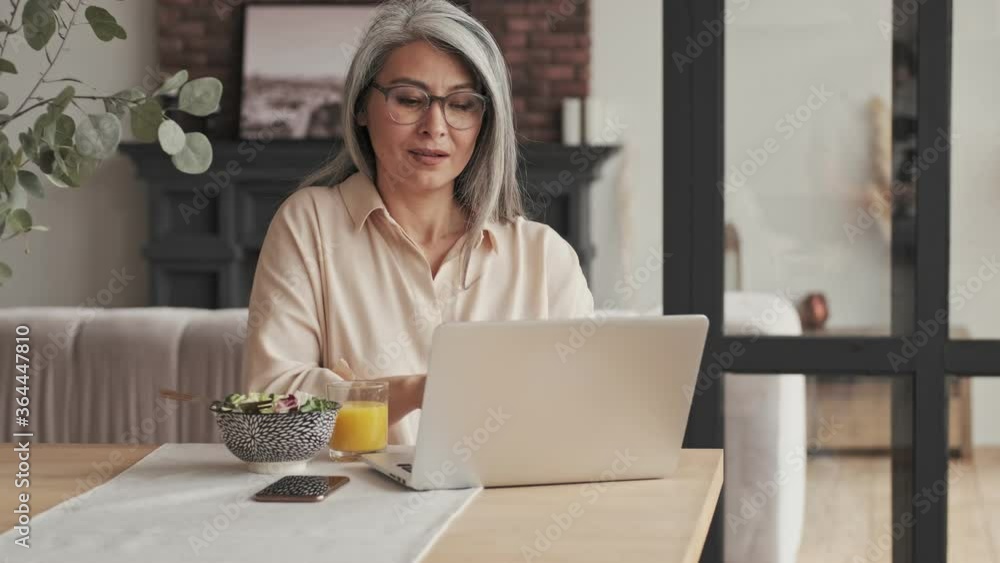 A pretty nice mature woman in eyeglasses is working with her silver laptop computer while eating food sitting at the table at home A pretty nice mature woman in eyeglasses is working with her silver laptop computer while eating food sitting at the table at home