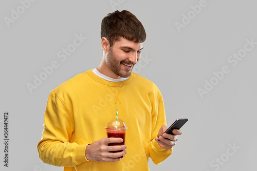 drinks and people concept - happy smiling young man with smartphone and tomato juice in takeaway plastic cup with paper straw over grey background