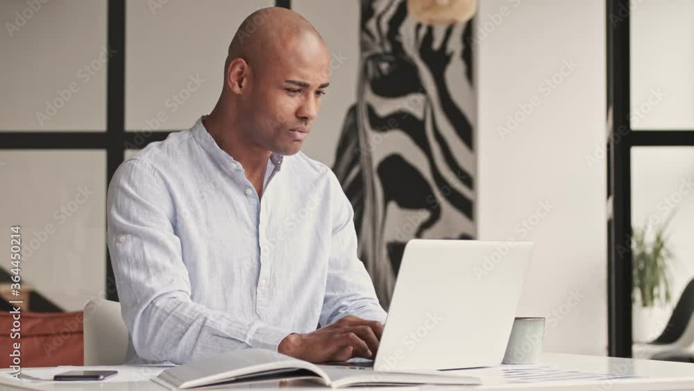 A confident focused african american man is working with laptop computer and smartphone sitting at table at home A confident focused african american man is working with laptop computer and smartphone sitting at table at home