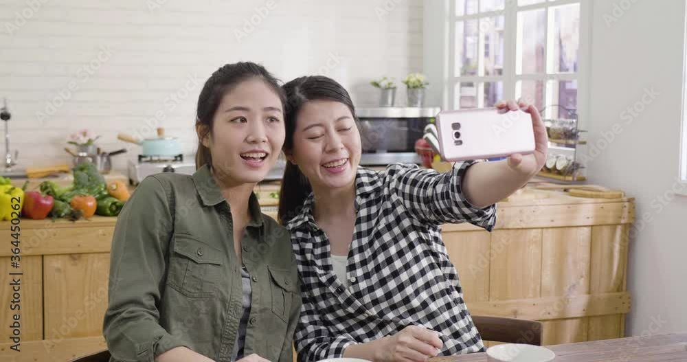 slow motion Two charming smiling korean woman friends taking selfie on smartphone at dining table. girls party in kitchen laughing and having fun. female roommates make self portrait at home slow motion Two charming smiling korean woman friends taking selfie on smartphone at dining table. girls party in kitchen laughing and having fun. female roommates make self portrait at home