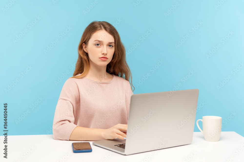 Naklejka premium Smart serious young pretty businesswoman in casual clothes looking at camera with responsible confident expression, working on laptop at home office. indoor studio shot isolated on blue background