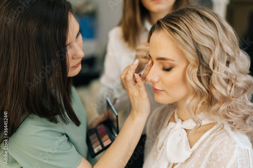 Professional make-up artist making face makeup while hairdresser making hair-do for young woman in beauty salon. Concept of backstage work.