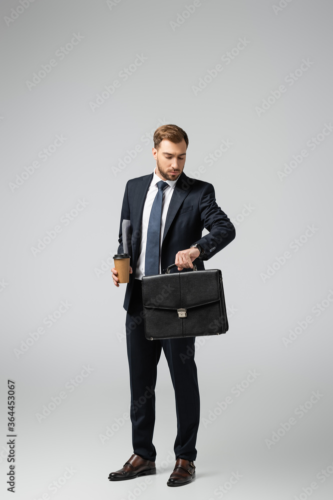 businessman with leather suitcase and paper cup looking at wristwatch isolated on grey businessman with leather suitcase and paper cup looking at wristwatch isolated on grey
