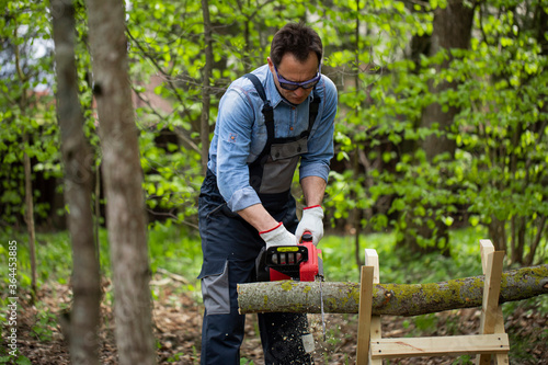 Close up view of woodcutter lumberman in working uniform sawing small tree trunk on sawhorse with electric saw chainsaw in forest on sawmill, outdoors. Wood work, handwork, wood cutting tools, timber