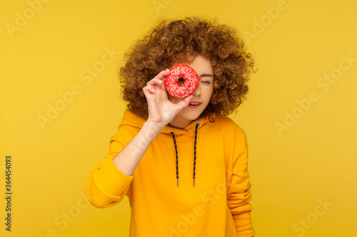 Portrait of curious curly-haired young woman in urban style hoodie looking through donut  having fun and playing with sugary food  appetizing doughnut. indoor studio shot isolated on yellow background