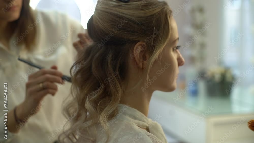 Back view of professional hairdresser making hairstyle young attractive woman and make-up artist applying makeup in beauty salon. Concept of beauty and fashion. Tracking shot in slow motion. Back view of professional hairdresser making hairstyle young attractive woman and make-up artist applying makeup in beauty salon. Concept of beauty and fashion. Tracking shot in slow motion.