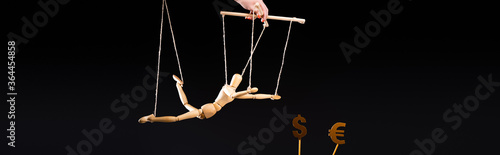 cropped view of puppeteer holding wooden marionette on strings near currency signs isolated on black, panoramic shot