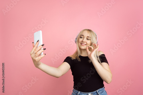 Taking selfie, vlog. Portrait of young woman with bright emotions on coral pink studio background. Blonde female model. Concept of human emotions, facial expression, sales, advertising, youth.
