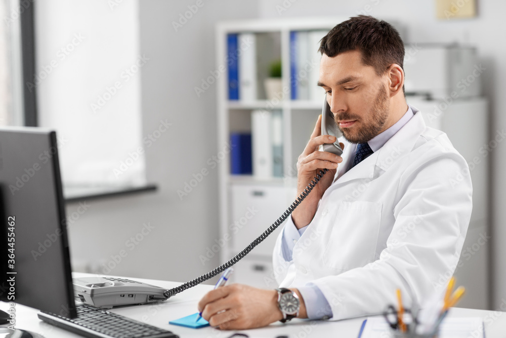 healthcare, medicine and people concept - male doctor with clipboard calling on desk phone at hospital