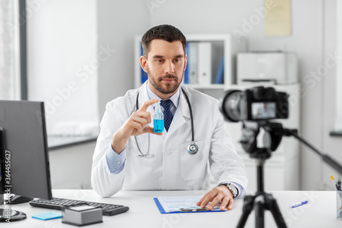 healthcare, medicine and blogging concept - male doctor with camera and hand sanitizer recording video blog at hospital