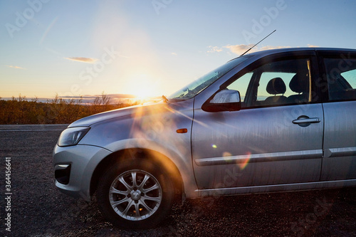Small car standing on the side of asphalt road and sunset or sunrise with rays of sun behind it. © keleny