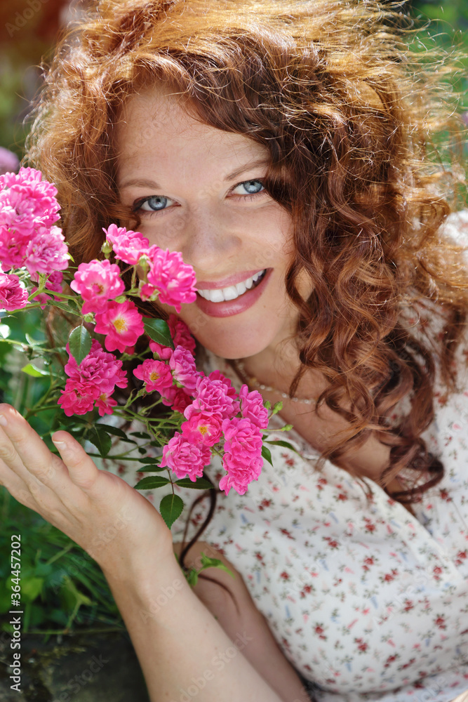 Naklejka premium jeune et jolie jeune femme rousse caucasienne romantique aux longs cheveux bouclés souriant à pleines dents dans une nature entourée de fleurs roses 