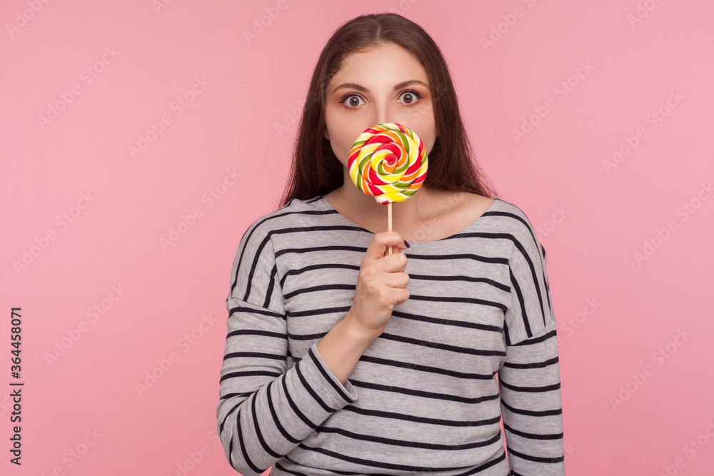 Portrait of amazed woman in striped sweatshirt licking lollipop, tasting sweet round rainbow candy with surprised expression, enjoying delicious flavor dessert. indoor studio shot, pink background