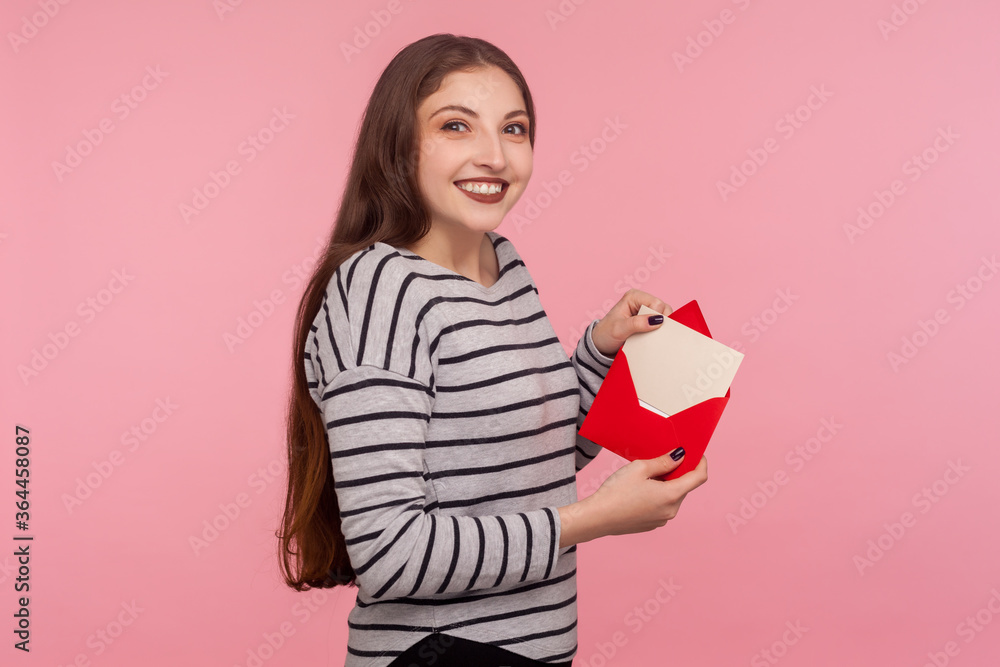 Portrait of charming woman in striped sweatshirt showing romantic letter in red envelope, holding love message and looking at camera with toothy smile. indoor studio shot isolated on pink background Portrait of charming woman in striped sweatshirt showing romantic letter in red envelope, holding love message and looking at camera with toothy smile. indoor studio shot isolated on pink background
