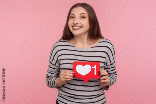 Click Like and follow my blog! Portrait of happy blogger woman in striped sweatshirt holding social media Heart button and looking at camera with toothy smile. studio shot isolated on pink background
