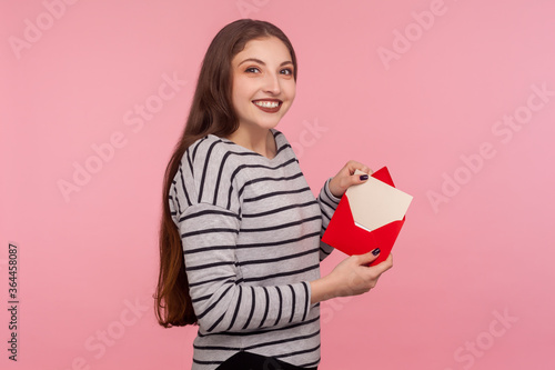 Portrait of charming woman in striped sweatshirt showing romantic letter in red envelope, holding love message and looking at camera with toothy smile. indoor studio shot isolated on pink background