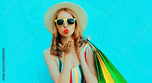 Portrait of woman with shopping bags blowing red lips sending sweet air kiss wearing colorful striped t-shirt, summer round hat on blue background