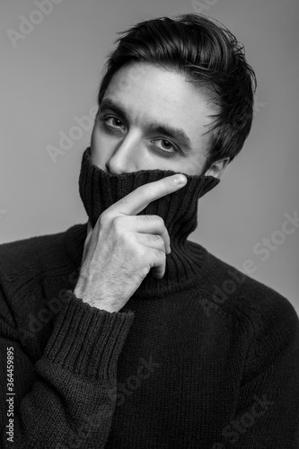 Portrait of stylish handsome young man in warm sweater, covering mouth with high collar and looking pensive thoughtful, male fashion, vogue concept. black and white, indoor studio shot isolated
