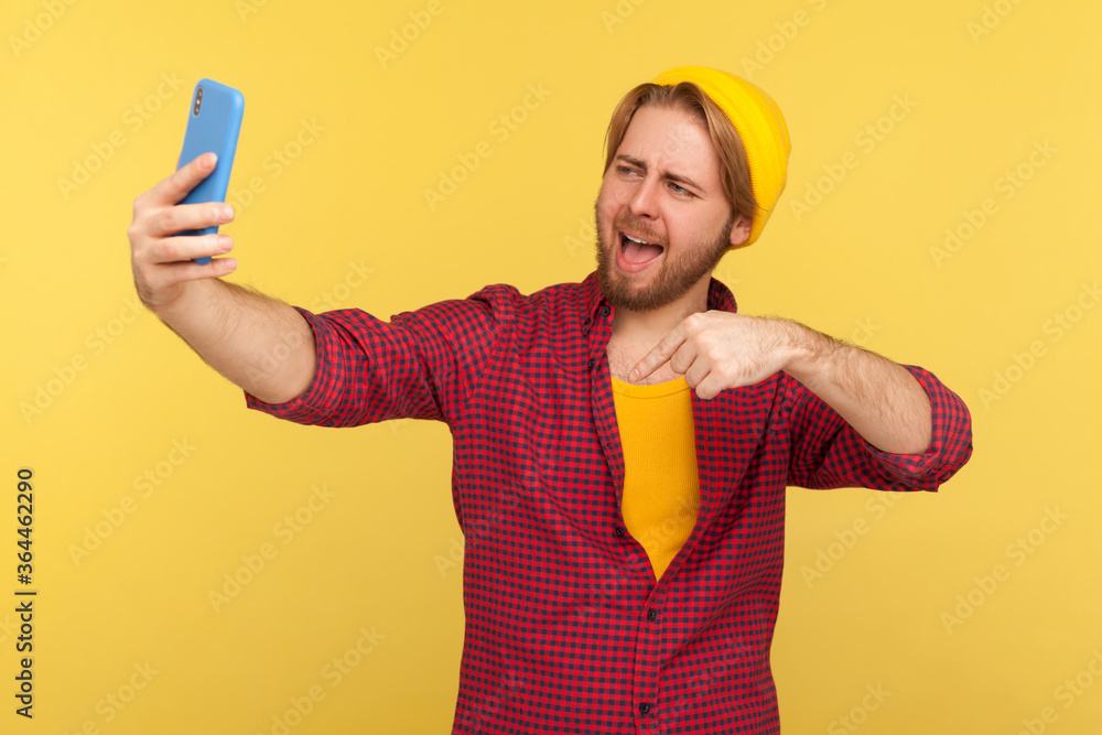 Optimistic trendy hipster guy in checkered shirt pointing finger down, click below, smiling talking video call or streaming on mobile phone, asking follow and subscribe his blog. studio shot isolated Optimistic trendy hipster guy in checkered shirt pointing finger down, click below, smiling talking video call or streaming on mobile phone, asking follow and subscribe his blog. studio shot isolated