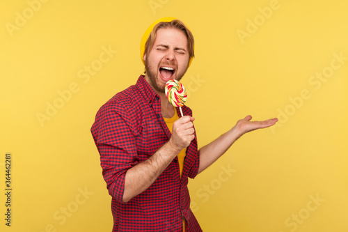 Billede på lærred Amazing hipster guy in beanie hat and checkered shirt screaming into lollipop like microphone, pretending to sing loudly in karaoke , having fun