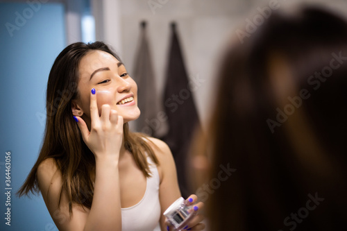Portrait of young asian woman applying moisturizer cream on her pretty face and look mirror.