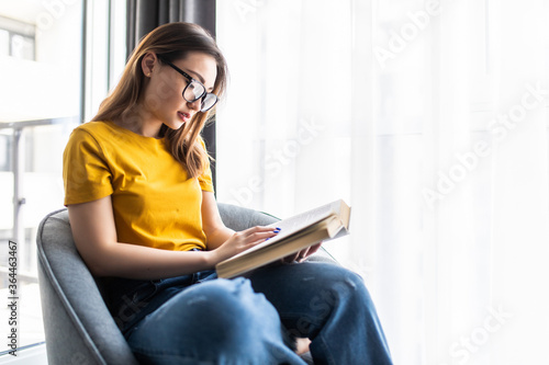 Asian young woman reading a book while sitting in modern armchair at home