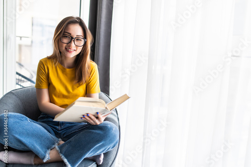 Asian young woman reading a book while sitting in modern armchair at home