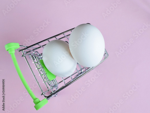 Two eggs in a small shopping cart. Ingredient for Easter cake. Mini trolley with eggs on a pink background. Selective focus. Copy space. photo