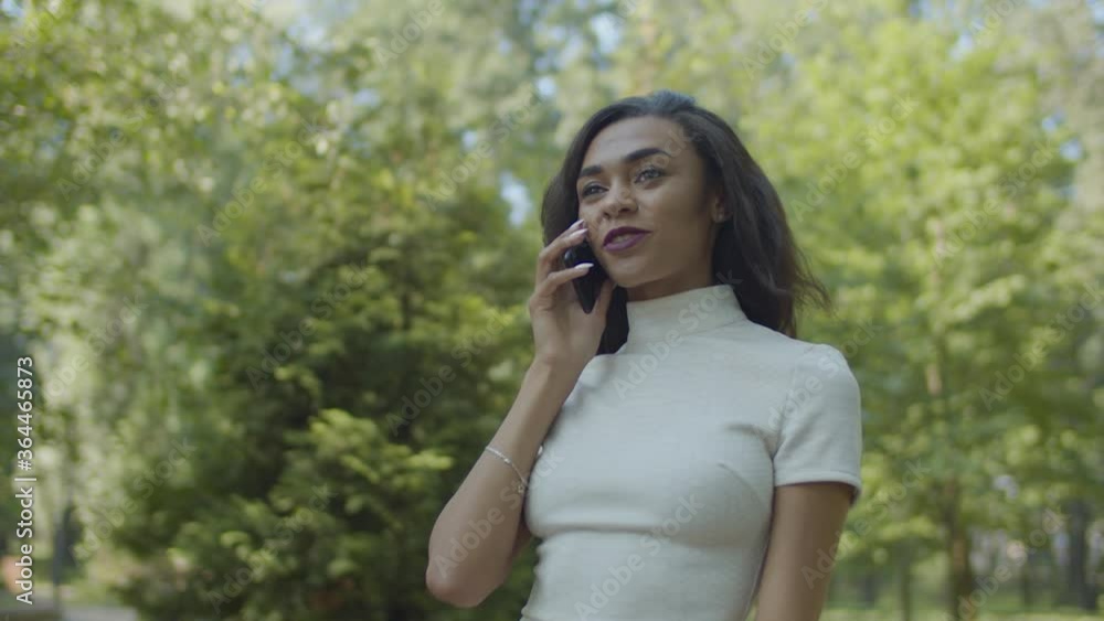 Portrait of elegant charming african american woman in stylish white dress talking on cellphone, walking along park alley on summer day, expressing confidence, beauty and attraction. Portrait of elegant charming african american woman in stylish white dress talking on cellphone, walking along park alley on summer day, expressing confidence, beauty and attraction.