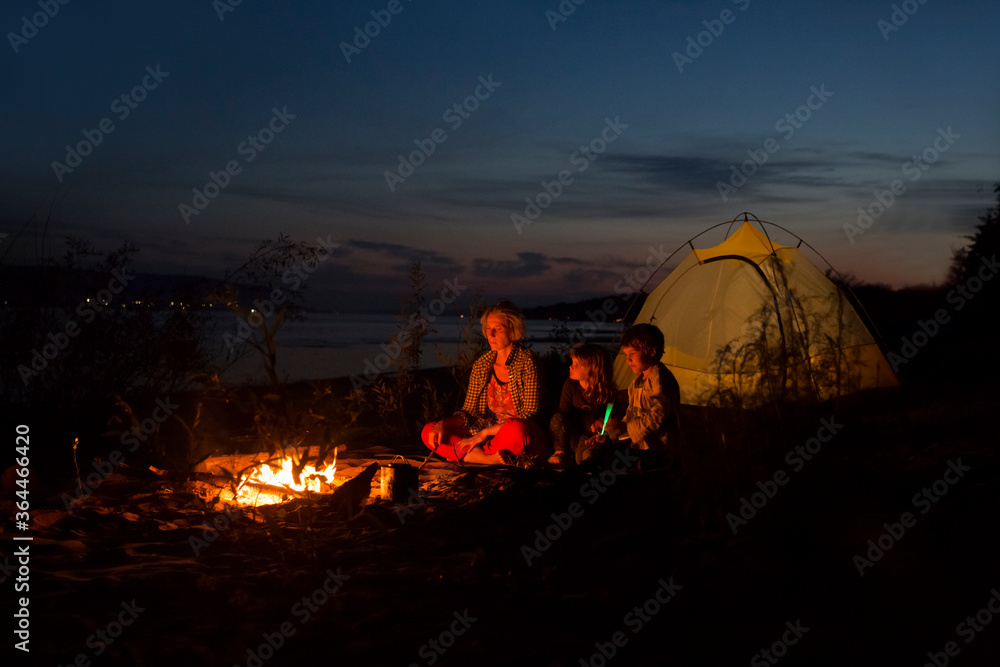 Family mom and two kids sitting together around campfire, enjoying fresh air near tent on the seashore. Tourism, camping concept. Staycations, hyper-local travel,  family outing, getaway.