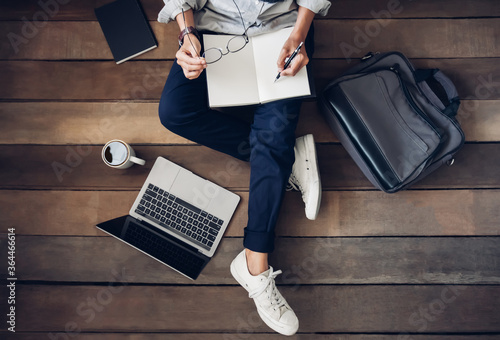 Man holding pencil writing notepad paper learning and research course tutoring education online on laptop with the coffee cup, briefcase, book on my desktop. Are sitting on the wooden floor. Top view photo