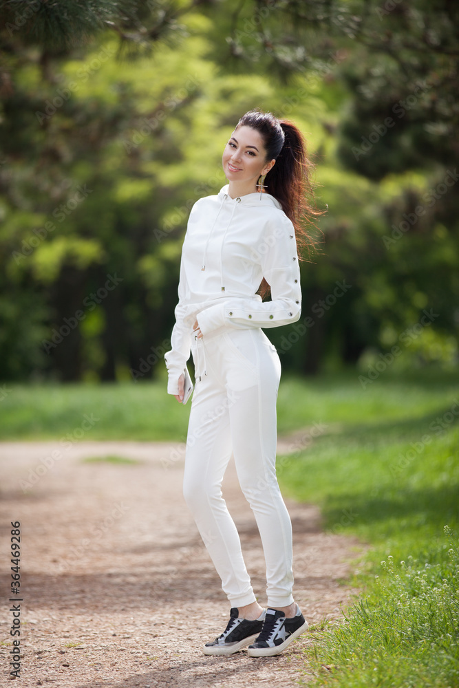 Young cute woman in white costume walking in the park. Beauty nature scene with colorful background, trees at summer season. Outdoor lifestyle. Trendy style Young cute woman in white costume walking in the park. Beauty nature scene with colorful background, trees at summer season. Outdoor lifestyle. Trendy style