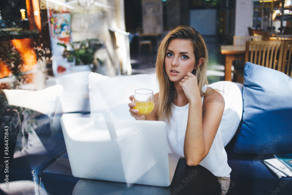 Young businesswoman with glass of juice in hand looking at camera while sitting with open net-book in cozy bar, beautiful female student enjoying cocktail during work on her portable laptop computer