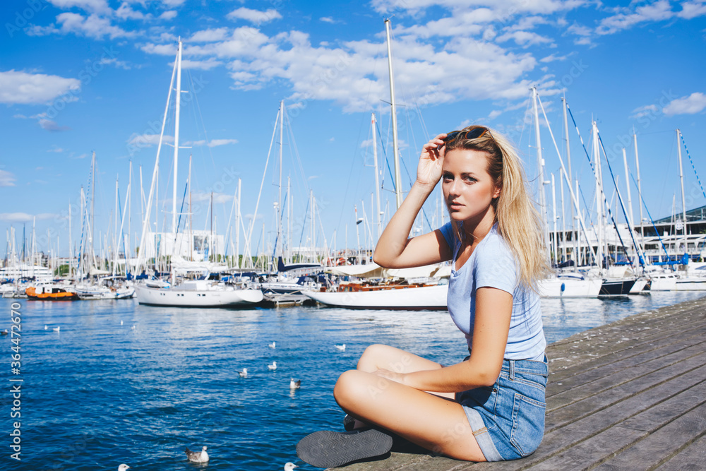 Young charming woman traveler posing while sitting on the wooden pier near yacht port during her summer weekend, gorgeous hipster girl looking at camera while enjoying good warm day in the fresh air