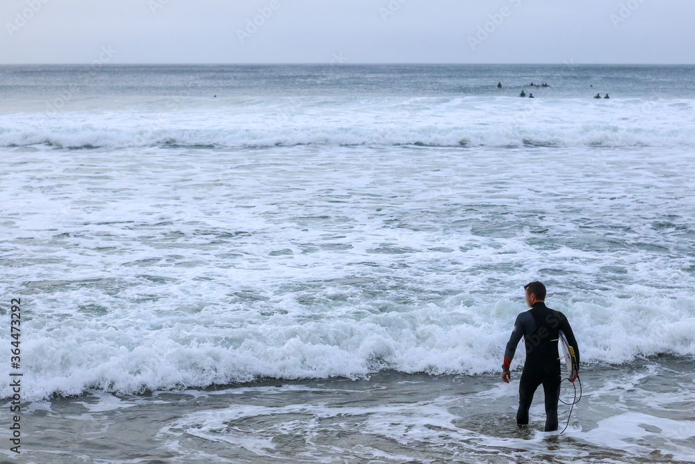 an unidentfiiable lone male surfer leading his surfboard through the waves off the beach on a rainy morning, as his friends surf in the background, Bell's beach, great ocean road, Victoria, Australia an unidentfiiable lone male surfer leading his surfboard through the waves off the beach on a rainy morning, as his friends surf in the background, Bell's beach, great ocean road, Victoria, Australia