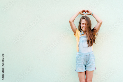 Portrait of a little girl child in a denim jumpsuit 9-13 years old on an empty wall, the girl shows a heart from her fingers