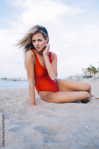 Portrait of seductive female dressed in trendy swimsuit sitting on the beach in cloudy summer day,caucasian blonde hair woman with perfect figure dressed in red swimwear rest after swimming in the sea