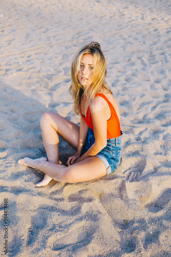 Portrait of a young beautiful woman dressed in stylish clothes sitting on the beach and enjoying leisure time after strolling by coastline, attractive female posing for the camera sitting on the sand