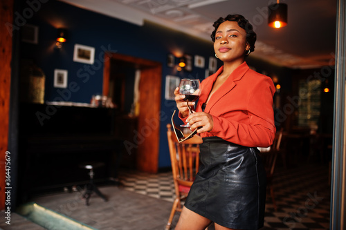 Portrait of african american woman, retro hairstyle with eyeglasses, wear orange jacket and leather skirt posing at restaurant with glass of wine.