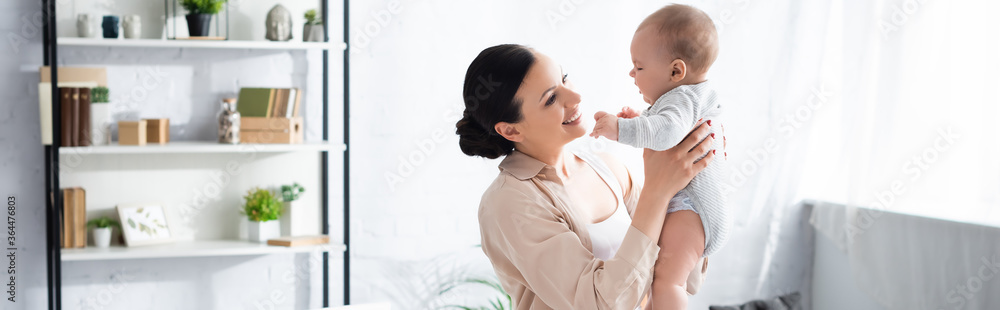 panoramic shot of cheerful mother holding in arms cute infant son in baby romper panoramic shot of cheerful mother holding in arms cute infant son in baby romper
