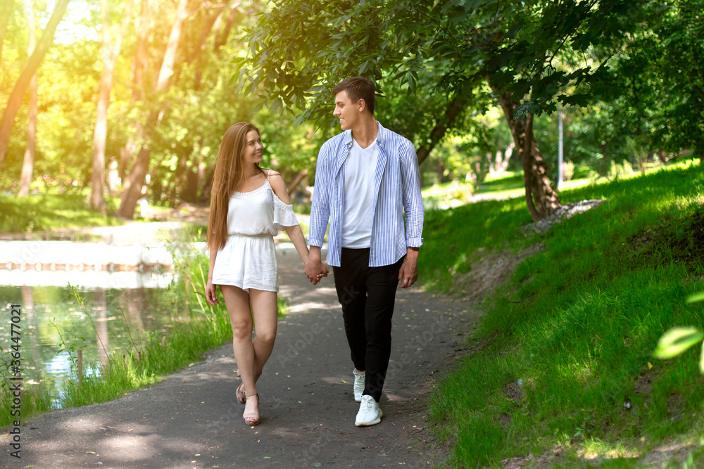 Affectionate millennial couple holding hands and walking along pathway in summer park Affectionate millennial couple holding hands and walking along pathway in summer park