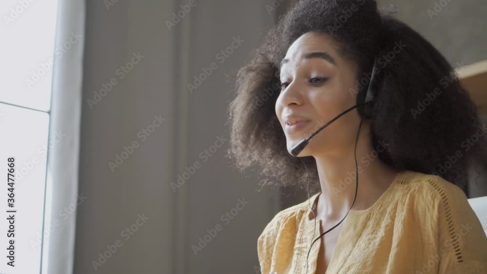 Smiling African American woman with headset using laptop, talking, working customer support service operator at hom\office, girl in headphones with microphone