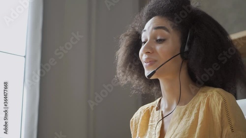 Smiling African American woman with headset using laptop, talking, working customer support service operator at homoffice, girl in headphones with microphone