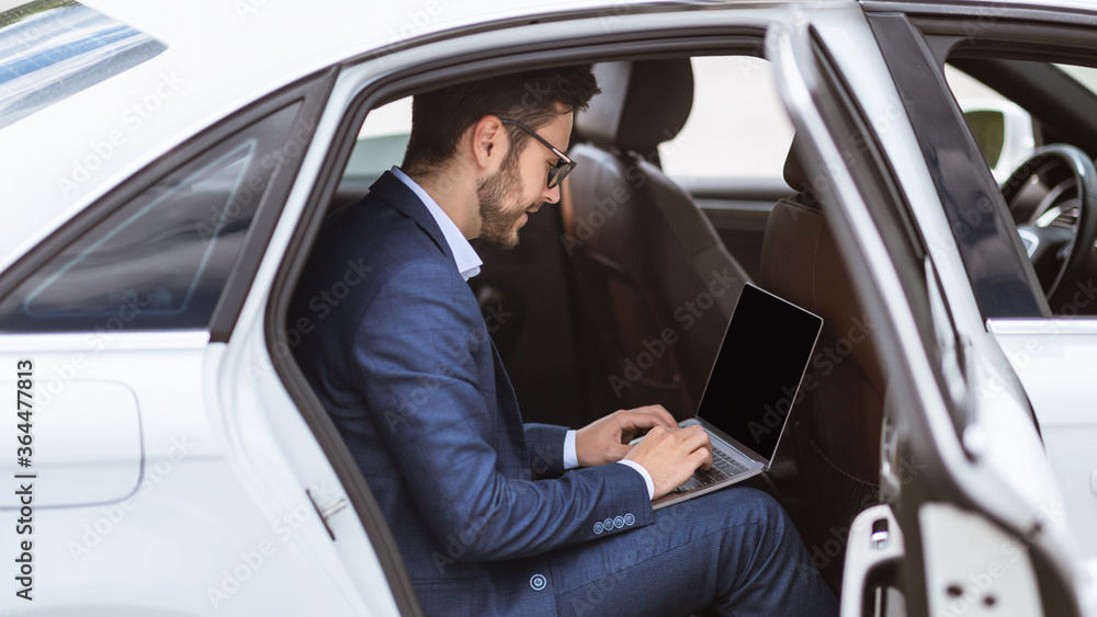 Young businessman using laptop computer with empty screen on back seat of his car, mockup for design