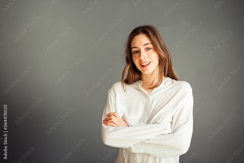 Fototapeta premium Young girl in a white shirt, hands up, on a gray background. Honest and emotional on the banner, billboard, billboard. No retouch. Without make-up.